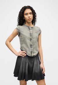 Γυναίκα με σγουρά μαλλιά που φοράει μια χωρίς μανίκια ξεθωριασμένη γκρι μπλούζα με κουμπιά και μια μαύρη τζιν φούστα με πιέτες, στέκεται με το ένα χέρι στη μέση της.