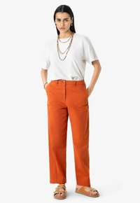 Pantalon en coton orange à coupe droite, t-shirt gris foncé rentré, colliers superposés et sandales beiges avec des détails tressés.