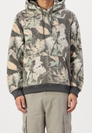 Man iklädd en luvtröja i fleece med gråa, beige och gröna abstrakta mönster samt beige cargobyxor med sidofickor.