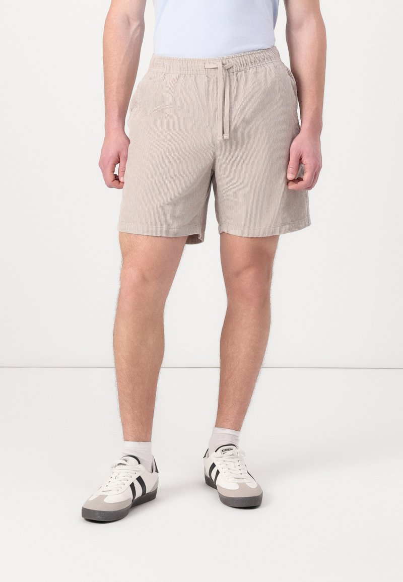 Pantalones cortos de color beige claro con un diseño de tejido acanalado, cintura elástica y cordón. Combinados con zapatillas deportivas blancas y grises.