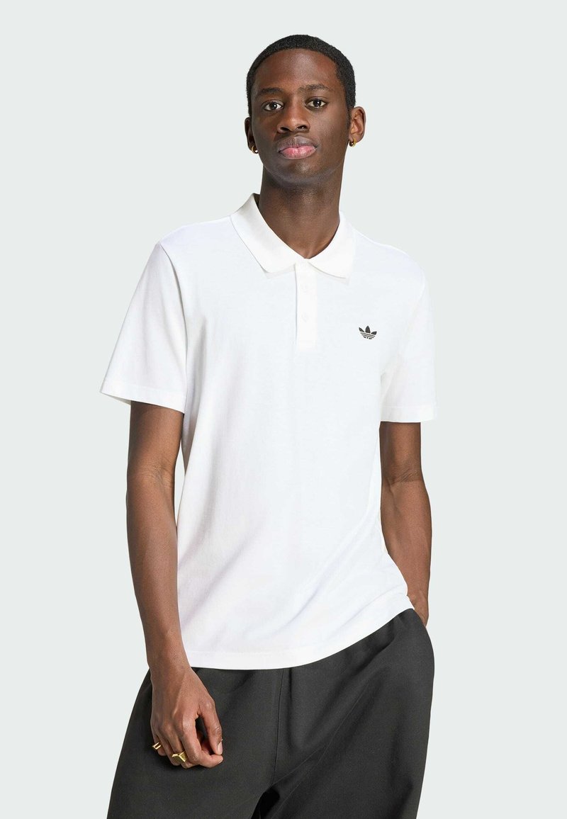 Junger Mann trägt weißes Adidas-Poloshirt mit Logo, schwarze Hose, steht mit einer Hand in der Tasche vor einfachem Hintergrund.