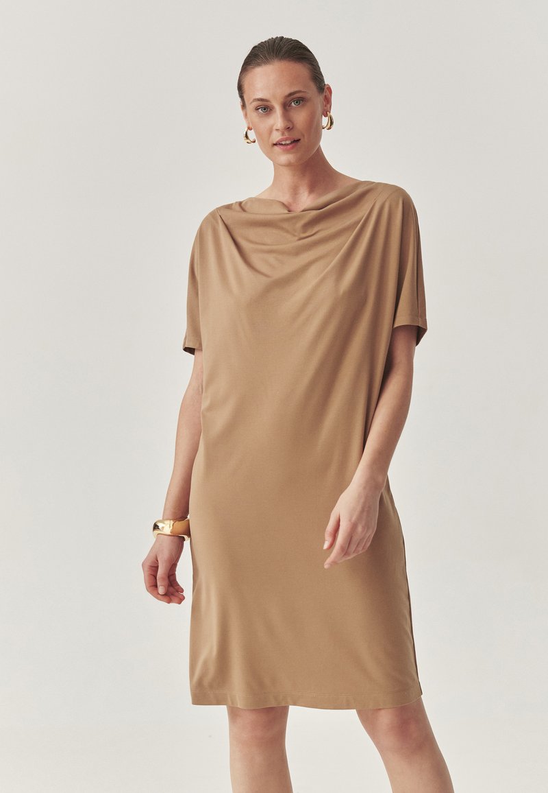 Cowl-neck, lyhythihainen mekko, joka on valmistettu sileästä, vaaleasta kankaasta. Polvipituinen, rento istuvuus, jossa on hienovarainen kiilto ja sivuhalkiot.