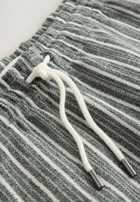 Grau-weiß gestreifter Stoff mit strukturierter Oberfläche. Verfügt über einen weißen Kordelzug mit schwarzen Metallspitzen und ein elastisches Taillenbund-Detail.
