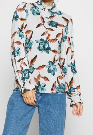 Kobieta nosząca kremową bluzkę z długim rękawem i golfem, z turkusowo-brązowym kwiatowym wzorem, w połączeniu z niebieskimi dżinsami.