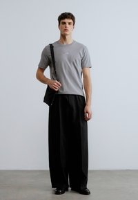 Γκρι κοντομάνικη μπλούζα με τυπωμένο λογότυπο, συνδυασμένη με φαρδιά μαύρη παντελόνια και μαύρη τσάντα χιαστί, πάνω σε τσιμεντένιο δάπεδο.