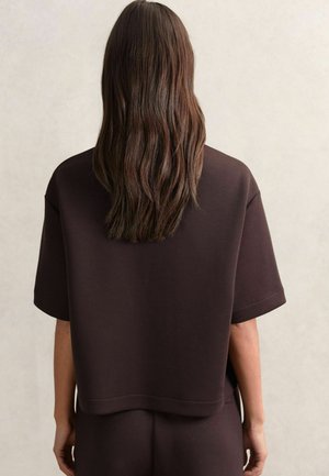 Persoană cu păr lung și brunet, purtând un top lejer de culoare maro închis cu mânecă scurtă și pantaloni asortați, stând cu spatele față de un fundal simplu.