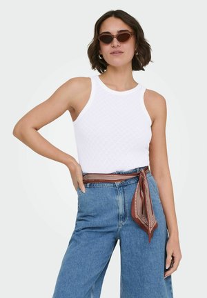 Mujer con top blanco sin mangas, jeans azules de cintura alta, cinturón con pañuelo marrón estampado y gafas de sol estilo ojo de gato, posando con una mano en la cadera.