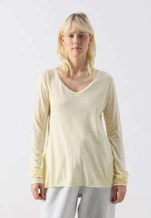 Mlajša ženska z ravnimi blond lasmi, oblečena v bledo rumeno majico z dolgimi rokavi in V-izrezom ter svetlo sive hlače, stoji ob preprostem ozadju.