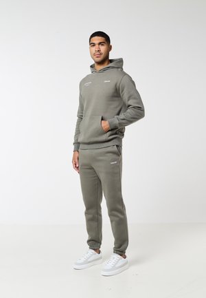 Hombre de pie con sudadera con capucha verde oliva y pantalones joggers a juego, con zapatillas blancas, posando frente a un fondo blanco liso.
