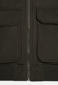 Mörk olivgrön jacka med dragkedja framtill, två stora fickor med lock, ribbad fåll och slät, strukturerad tyg.