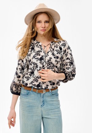 Młoda kobieta w beżowym kapeluszu, kwiecistej bluzce z długim rękawem, brązowym pasku i jasnoniebieskich dżinsach na białym tle.