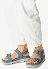 Blå denimsandaler med flerbandsdesign, prydda med dekorativa stenar och en ljus färgad plattformssula. Vita byxor synliga.
