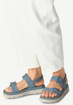 Blauwe denim sandalen met een multi-band ontwerp, voorzien van decoratieve stenen accenten en een lichtgekleurde platformzool. Witte broek zichtbaar.