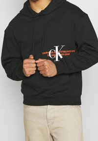 Černá mikina s kapucí a přední kapsou kangaroo, na hrudi s potiskem bílého a oranžového textu. Vyrobená z bavlněné směsi.