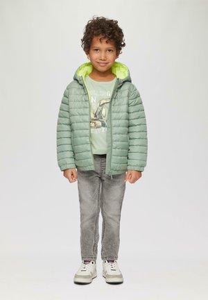 Hellgrüner Pufferjacke mit einer leuchtend gelben Kapuze, gestepptes Design, kombiniert mit einem hellgrünen T-Shirt und grauen Jeans sowie weißen Sneakers.