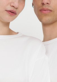 Κοντινό πλάνο δύο ατόμων που φορούν λευκά πουκάμισα, δείχνοντας το κάτω μέρος του προσώπου και την περιοχή του λαιμού· ένα άτομο έχει σκουλαρίκι στο αριστερό αυτί.
