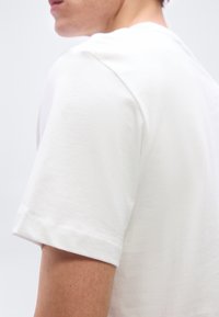 Κοντινό πλάνο ατόμου που φοράει απλό λευκό κοντομάνικο μπλουζάκι, δείχνοντας τον ώμο, το πάνω μέρος του μπράτσου και μέρος του προφίλ του προσώπου.