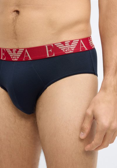 Cueca masculina em tecido azul marinho com cós elástico vermelho apresentando um logótipo em texto claro e emblema de águia, desenhada para um ajuste justo.