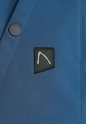 Sininen kangastakki, jossa on musta neliön muotoinen päällyste, jossa on valkoinen abstractti kuvio, sekä pyöreä musta nappi, jossa lukee "CHASIN."