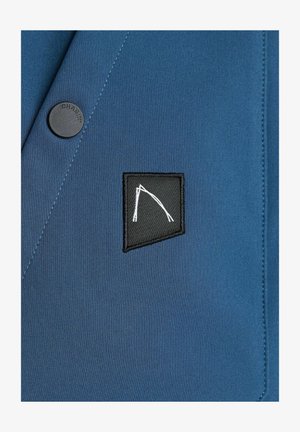 Niebieska marynarka z tkaniny z czarną kwadratową naszywką z białym abstrakcyjnym wzorem oraz okrągłym czarnym guzikiem z napisem "CHASIN."