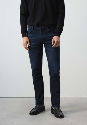 Hombre con vaqueros ajustados azul oscuro, camisa negra de manga larga y mocasines negros de cuero, de pie sobre un suelo gris claro.