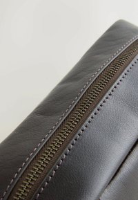 Secțiune a unei genți din piele întunecată, cu un fermoar proeminent cu dinți aurii și cusături contrastante de-a lungul marginii.