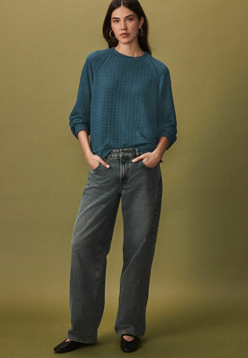 Teal textuur trui met een ronde hals, gecombineerd met hoge, losse grijze jeans en zwarte platte schoenen. Basis ringoorbellen.