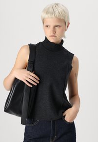 Top sin mangas de color gris oscuro con cuello de tortuga de canalé; textura suave. La modelo sostiene un bolso de hombro de cuero negro con una correa ancha.