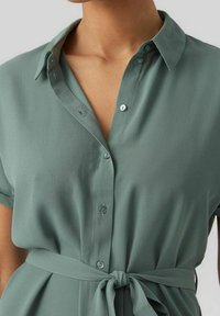 Vaaleanvihreä pusero kevyestä kankaasta, jossa on kaulus, lyhyet hihat, napitus ja solmittu vyötärönauha.