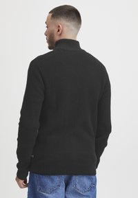 Svart strikkegenser med høy krage, med et teksturert m�ønster og ribbede mansjetter. Bært sammen med blå denimjeans, sett bakfra.