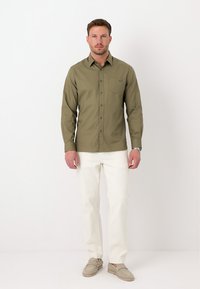 Homme debout sur fond blanc, portant une chemise boutonnée vert olive, un pantalon blanc, des mocassins beiges et une montre-bracelet.