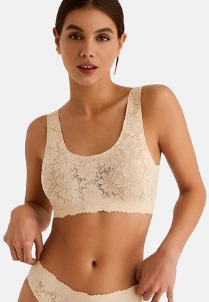 Femme portant un soutien-gorge en dentelle florale beige et une lingerie assortie, posant avec la main ajustant la taille de la lingerie.