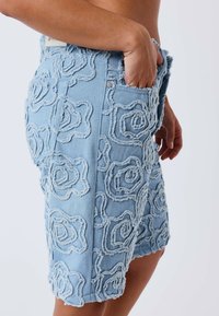 Short en denim bleu clair avec détail d'appliqué floral texturé, porté par une personne avec une main dans la poche avant sur un fond uni.