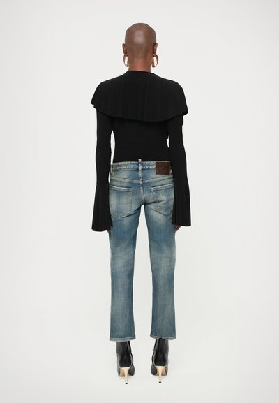 Άτομο με ξυρισμένο κεφάλι φοράει μεγάλα σκουλαρίκια κρίκους, μαύρο μακρυμάνικο τοπ με λεπτομέρεια κάπας, μπλε τζιν και μαύρες μπότες με ψηλό τακούνι, κοιτάζοντας από πίσω.