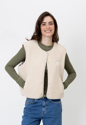 Donna sorridente con capelli castani indossa un gilet in pile beige sopra una maglia verde a maniche lunghe e jeans blu, in piedi con le mani nelle tasche del gilet.