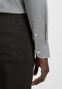 Black striped dress shirt with a buttoned cuff, made of fabric. Paired with black pants featuring subtle stitching and a rear pocket.