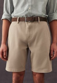 Pantalones cortos beige de una tela texturizada con corte recto, llevados con un cinturón marrón que presenta una hebilla metálica cuadrada. Camisa de rayas verticales arriba.