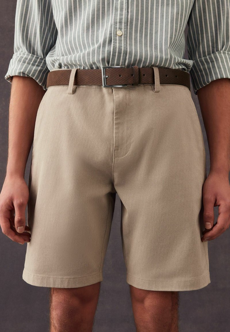 Pantalones cortos beige de una tela texturizada con corte recto, llevados con un cinturón marrón que presenta una hebilla metálica cuadrada. Camisa de rayas verticales arriba.