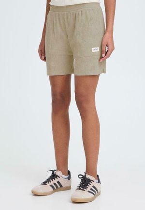 Osoba w beżowych szortach o fakturze z naszywaną kieszenią i beżowych tenisówkach z czarnymi paskami, stojąca na jasnym tle.