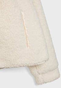 Kremno obarvana flis jakna z teksturirano, puhasto površino. Ima skrite stranske žepe in sproščen silhuet z šivanimi robovi.