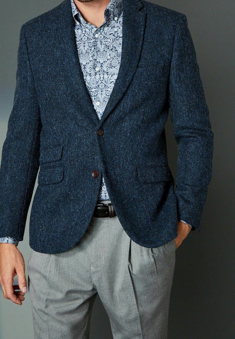 Blazer azul marino de espina de pescado con dos bolsillos delanteros, botones marrones, combinado con una camisa de estampado floral y pantalones gris claro.