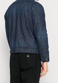 Jeansjacka i mörkblått med klassisk design, med knäppning i ärmsluten, sidofickor och en sydd fåll. Den kombineras med svarta byxor.