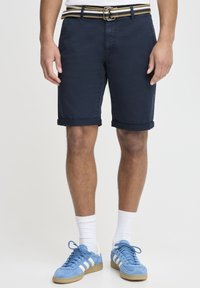 Mornarsko plave pamučne kratke hlače s prevrnutim manžetama, u paru s prugastim pojasom. Nosene s plavim tenisicama i bijelim čarapama, pokazuju opušteni, sportski stil.