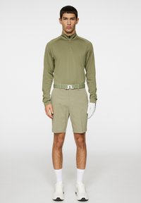 Vaaleanoliivinvihreä pitkähihainen vetoketjullinen paita, matching shortsit, valkoinen vyö korostuksella, valkoinen golf-käsine ja valkoiset urheilukengät.