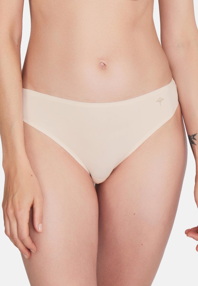 Femme portant un sous-vêtement style bikini beige clair uni avec un petit logo sur la hanche droite, montrée de la taille aux cuisses.
