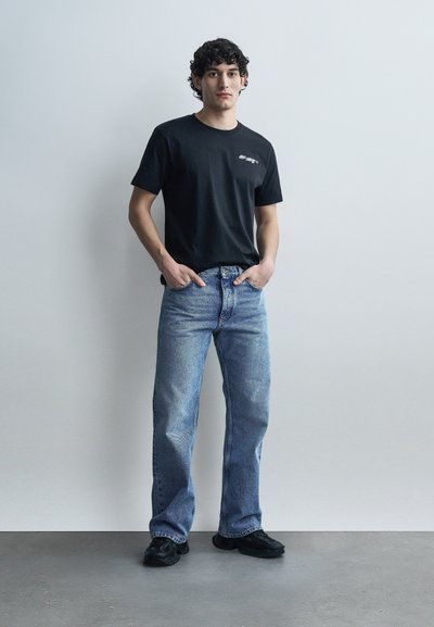 Jeune homme aux cheveux bouclés debout contre un mur uni, portant un t-shirt graphique noir, un jean bleu et des baskets noires, les mains dans les poches.