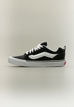 Zapatilla deportiva negra de corte bajo con cordones blancos, una franja lateral blanca y una suela blanca gruesa, vista de perfil sobre un fondo liso.