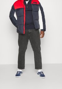 Muž v červené, námořnické a bílé péřové bundě přes námořnické tričko, černé džíny, bílé ponožky Adidas a modré tenisky Adidas, stojící s rukama v kapsách.