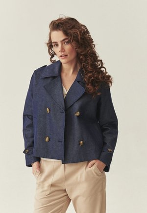 Žena s dlhými kučeravými vlasmi, oblečená v tmavomodrom dvojsrdečnom saku a béžových nohaviciach, ruky v vreckách, neutrálne pozadie.