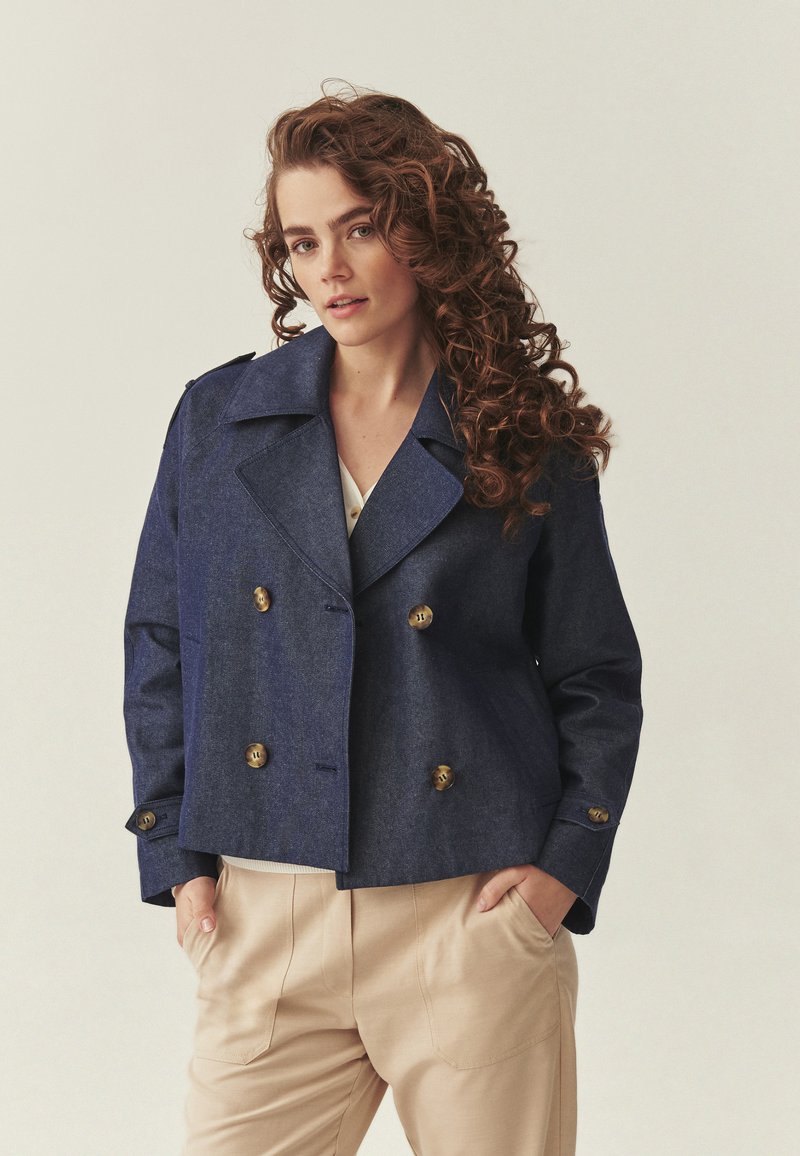 Femme aux cheveux longs et bouclés portant une veste bleu foncé à double boutonnage et un pantalon beige, les mains dans les poches, fond neutre.